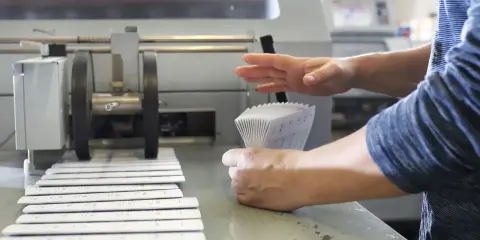 Mains manipulant une pile de documents imprimés pour le façonnage imprimerie, à côté d'une machine.