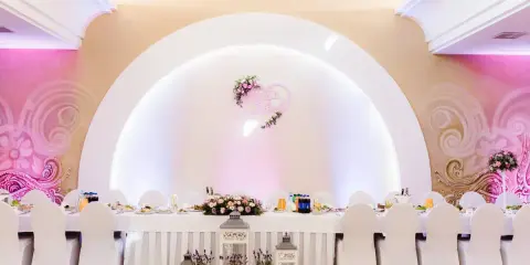 Elegancka sala weselna z białymi pokrowcami na krzesłach i serwowanym stołem. Serce z imionami pary młodej zdobi ścianę, tworząc atmosferę wesela z klasą.