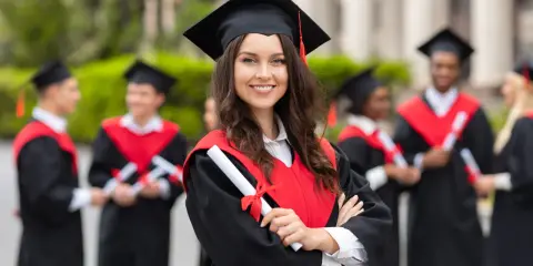 Uśmiechnięta absolwentka z dyplomem w ręku, w tle inni studenci po ang.