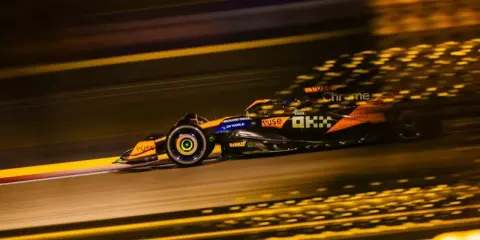 Un coche de **fórmula 1** naranja y negro acelera en la noche, dejando estelas de luz dorada.
