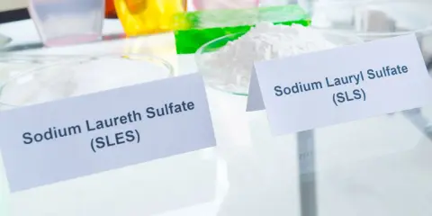 Zwei Schalen mit weißen Pulvern, beschriftet mit "Sodium Laureth Sulfate (SLES)" und "Sodium Lauryl Sulfate (SLS)". Diese Inhaltsstoffe sind oft in SLS-Shampoos zu finden.