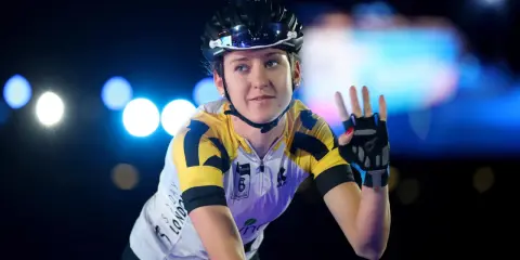 Lea Lin Teutenberg winkt mit der Hand, trägt Radsportkleidung und einen Helm. Blaue und weiße Lichter im Hintergrund.