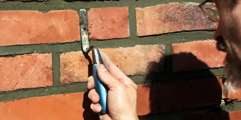 Hand mit Kelle bessert Zementfugen einer Ziegelwand aus.