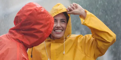Zwei lachende Personen im Regen, eine in Gelb, die andere in Rot. Die gelbe Regenjacke ist im Fokus.