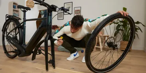 Mężczyzna klęczy przy rowerze, używając małego żółtego narzędzia, by pokazać, jak zdjąć oponę z roweru bez łyżki.