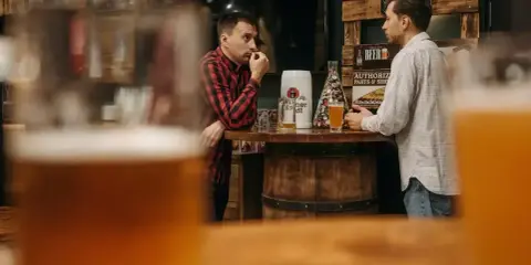 Dwóch mężczyzn rozmawia przy barze, zastanawiając się, ile gram alkoholu ma piwo. Na blacie stoją kufle i piwo.
