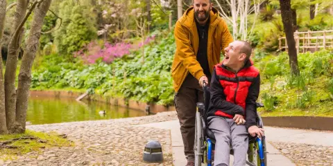 Mężczyzna pcha wózek z uśmiechniętym chłopcem. W tle piękny park z jeziorkiem. Pokazuje, że niepełnosprawność sprzężona nie ogranicza radości życia.
