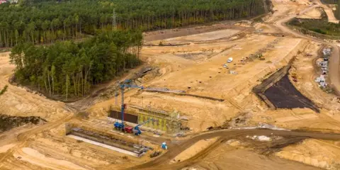 Ogromna budowa dróg w Polsce. Teren prac budowlanych z maszynami, dźwigami i przygotowywaną nawierzchnią, otoczony lasem.