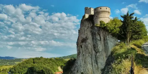 La fortezza di San Leo domina un picco roccioso, con un borgo sottostante e un cielo azzurro punteggiato di nuvole.