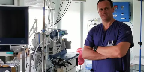 Lekarz w niebieskim uniformie stoi na tle aparatury medycznej. Jeśli masz ból jądra, to właśnie do takiego specjalisty powinieneś się zgłosić.
