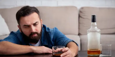 Mężczyzna z zarostem patrzy na szklankę z alkoholem, widoczne objawy uzależnienia. Obok pusta butelka i kolejna szklanka.