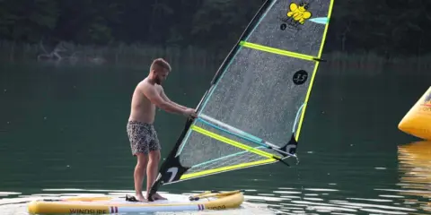 Mężczyzna na desce z żaglem, gotowy do wind sup. Na żaglu pszczółka i napis "3.5".