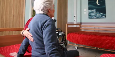 Anziana in stanza, forse affetta da sindrome da ospedalizzazione, con un'altra persona che le appoggia una mano sulla spalla. Sulla parete un quadro con luna e nuvole.