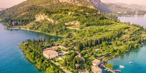Punta San Vigilio: eine Villa mit Gärten und Zypressen am türkisfarbenen Gardasee, umgeben von grünen Hügeln.