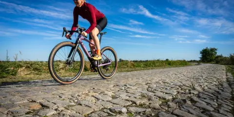 Kobieta w kasku i okularach jedzie na rowerze BMC URS LT po brukowanej drodze.