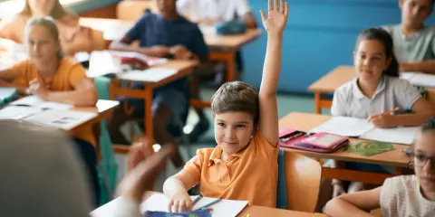 Chłopiec z podniesioną ręką czeka na ocenianie odpowiedzi. Inne dzieci pracują przy ławkach.