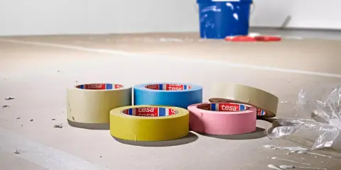 Fem rullar maskeringstejp i olika färger ligger på ett golv som täckts med papper. En blå hink och en röd roller syns i bakgrunden.