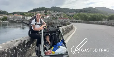Frau mit Rucksack und Kinderwagen auf einer Brücke, bereit für eine abc tour. Im Hintergrund eine Stadt und Berge.