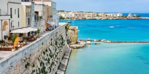 Vista del lungomare di Otranto centro, con edifici bianchi, un ristorante all'aperto e il mare azzurro cristallino.