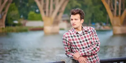 Młody mężczyzna w kratowanej koszuli i dżinsach opiera się o balustradę nad rzeką. W tle widać most.