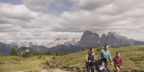 Famiglia in escursione sull'Alpe di Siusi, con bambini e passeggino, sullo sfondo le maestose vette dolomitiche.