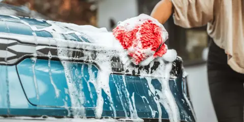 Mano con guante rojo y esponja enjabonada limpiando la carrocería del coche azul.