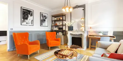 Salon aux fauteuils orange vif, cheminée ornée, et un jeu d'échecs sur table basse, évoquant un mouvement baroque moderne.