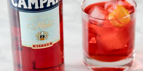 Butelka Campari i szklanka z czerwonym drinkiem, lodem i skórką pomarańczy. Idealny aperitif, który może być bazą dla drinków z Aperolem.