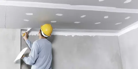 Pracownik w żółtym kasku nakłada masę szpachlową na sufit, przygotowując go do montażu. To kluczowy etap, by wiedzieć, jak przykleić styropian do sufitu.