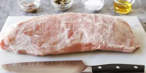Un pezzo di girello di vitello crudo, pronto per essere cucinato, con coltello e ciotoline di condimenti sullo sfondo.