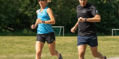 Two runners, a woman in blue and a man in black, run on a gravel path with a grassy field and trees in the background.