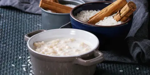 Gotowy do podania ryż na mleku w ceramicznej miseczce, obok miseczki z ryżem i laskami cynamonu.