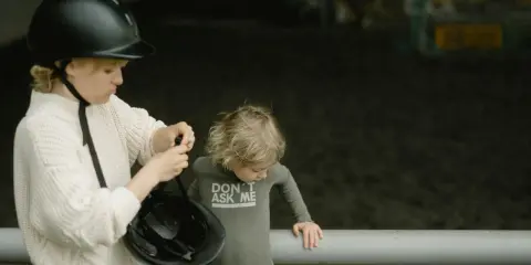 Starsza dziewczynka w kasku pokazuje młodszemu dziecku, jak zmierzyć głowę na kask, przygotowując się do jazdy konnej.