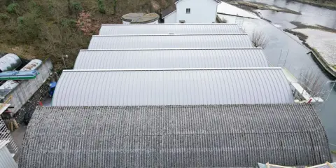 Reihen von Sheddach-Konstruktionen mit Metall- und Wellplatten. Ein Fluss und ein Gebäude sind im Hintergrund zu sehen.
