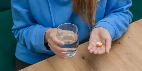 Kobieta w niebieskiej bluzie trzyma szklankę wody i kilka tabletek na dłoni.