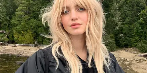 Blonde Frau mit lässigem Wolf Cut stylen, blauen Augen und einem schwarzen Jackett vor einer Waldkulisse.