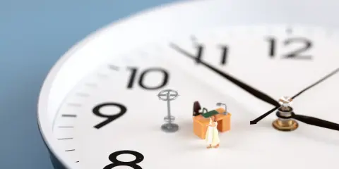 Figurka kobiety w sukience stoi przy miniaturowym biurku, symbolizując pracę w godzinach nadliczbowych. Zegar wskazuje późną godzinę.
