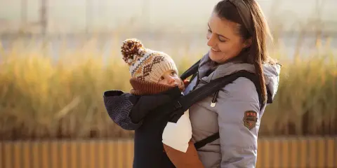 Uśmiechnięta mama z dzieckiem w nosidełku dla dziecka w góry. Maluch w czapce z pomponem i brązowym szaliku.