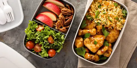 Dwa pojemniki z jedzeniem: jeden z kurczakiem w sosie, ryżem i natką, drugi z sałatką, jabłkami i orzechami. Idealne fit przepisy airfryer na lunch.