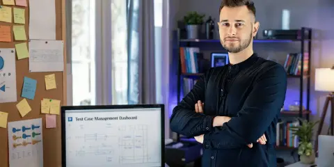 Mężczyzna w czarnej koszuli stoi obok biurka z monitorem wyświetlającym dashboard zarządzania testami. Tablica z notatkami i schematami podkreśla jego zaangażowanie w **quality engineering**.