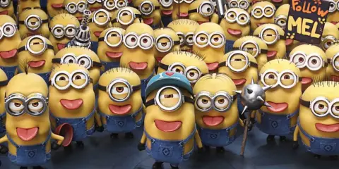 Grupa wesołych, żółtych minionków postacie w niebieskich ogrodniczkach, jeden z nich trzyma tabliczkę "Follow MEL".
