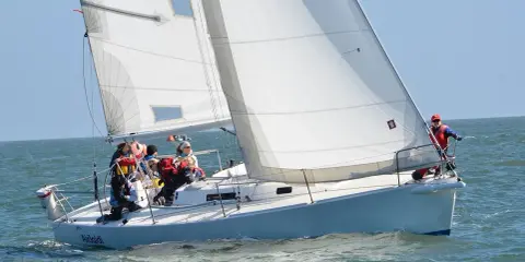 Un voilier blanc navigue sur une mer agitée, avec plusieurs personnes à bord. Le soleil brille, les voiles sont gonflées par le vent.