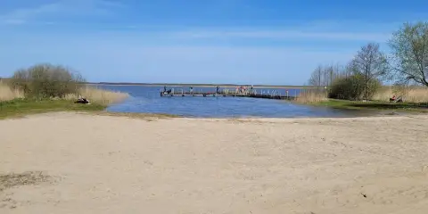 Plaża nad jeziorem Wicko, z pomostem, na którym stoją ludzie. W oddali widać trzciny i drzewa.