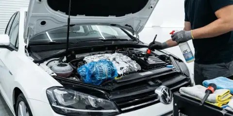 Persona con guantes rocía producto para limpiar motor coche. Usa cepillo para detallar el compartimento del motor cubierto de espuma.