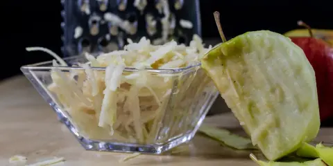 Frisch geriebener Apfel in einer Glasschale, daneben ein Apfelstück und eine Reibe.
