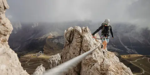 Kobieta w kasku i uprzęży pokonuje via ferrata, czyli ubezpieczoną trasę w górach. Widok zapiera dech w piersiach.