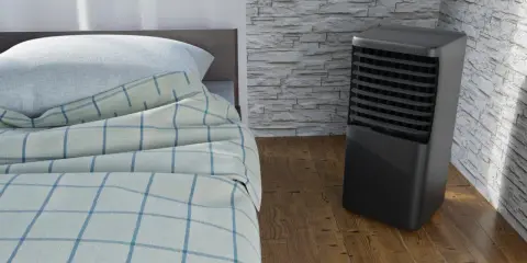 Un aire acondicionado portátil junto a una cama con sábanas a cuadros. ¿Qué es mejor, aire acondicionado portátil o fijo?
