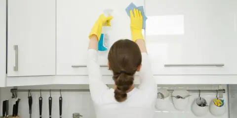 Mujer con guantes amarillos realiza limpieza armarios cocina, rociando y limpiando con paño azul.