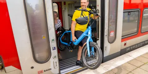 Mann mit blauem E-Bike steigt in den Zug. Perfekt, um das E-Bike im Zug mitzunehmen und die Reise fortzusetzen.