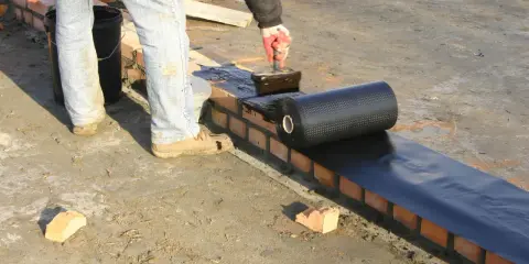 Pracownik nakłada masę asfaltowo kauczukową na mur ceglany, przygotowując podkład pod hydroizolację.